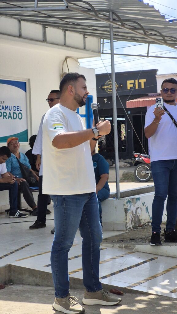 Alcalde habla con un micrófono durante un acto comunitario al aire libre frente a la sede del Camu del Prado, mientras asistentes observan y registran la actividad.