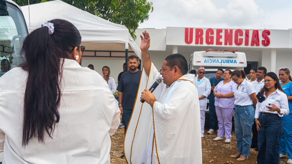 Sacerdote dirige la ceremonia de bendición de la ambulancia frente al área de Urgencias, mientras personal de salud y miembros de la comunidad observan.