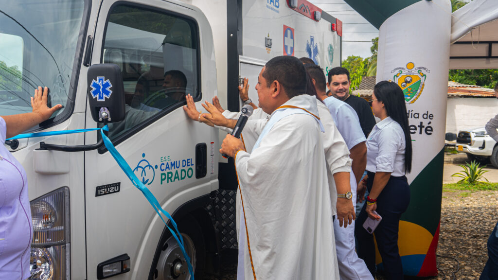 Un sacerdote bendice una nueva ambulancia de la E.S.E. CAMU del Prado durante un acto institucional, acompañado por funcionarios y asistentes. El vehículo está decorado con una cinta azul y, al fondo, se observa un elemento con el escudo de la Alcaldía de Cereté.