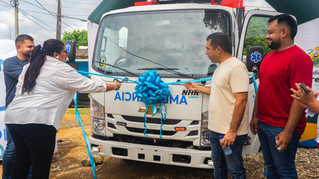 La gerente corta la cinta azul frente a una nueva ambulancia decorada con un gran lazo, mientras varios asistentes y el alcalde acompañan el acto oficial de entrega del vehículo.